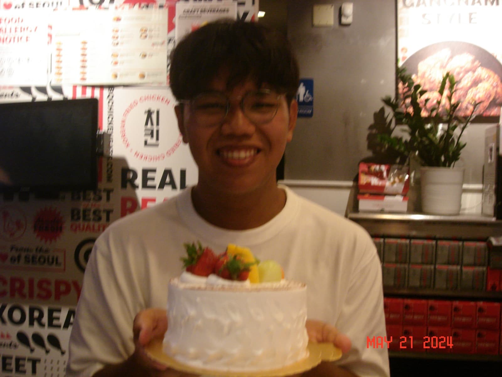 Erik grinning while holding a fruit-topped cream cake in a Korean fried chicken restaurant.
